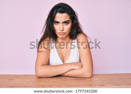 Brunette teenager girl wearing casual clothes sitting on the table skeptic and nervous, disapproving expression on face with crossed arms. negative person. 