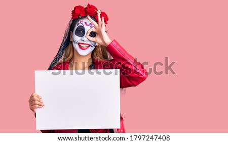Woman wearing day of the dead costume holding blank empty banner smiling happy doing ok sign with hand on eye looking through fingers 