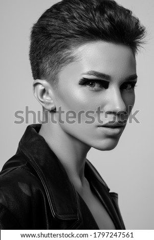 Beautiful confident woman with strong face, bold eyeliner makeup, wearing black trendy leather jacket, posing on grey background. Black and white, monochrome close up studio fashion portrait Royalty-Free Stock Photo #1797247561