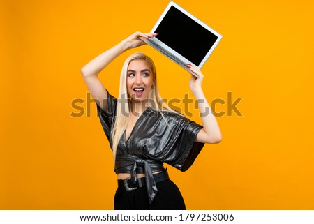 smiling charming blond girl holds a laptop screen blank over her head against a yellow wall