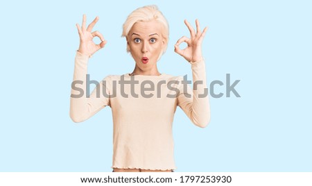 Young blonde woman wearing casual clothes looking surprised and shocked doing ok approval symbol with fingers. crazy expression 