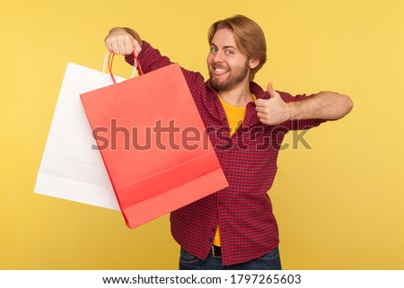 Satisfied shopper, handsome stylish guy in checkered shirt holding shopping bags blank mock up package, looking at camera with thumbs up like gesture. indoor studio shot isolated on yellow background