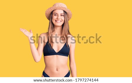Young beautiful girl wearing bikini and hat smiling cheerful presenting and pointing with palm of hand looking at the camera. 