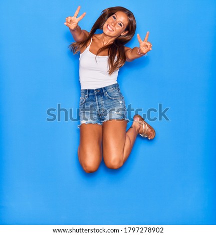 Young beautiful hispanic woman wearing casual clothes smiling happy. Jumping with smile on face doing victory sign over isolated blue background.
