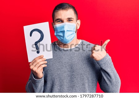 Young handsome man wearing medical mask holding question mark smiling happy and positive, thumb up doing excellent and approval sign 