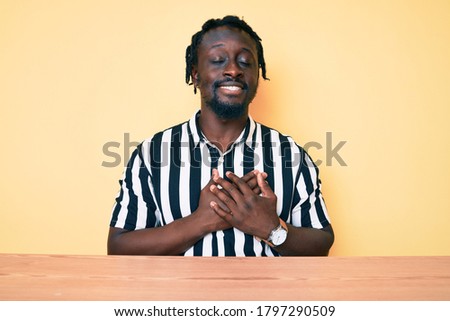 Young african american man with braids wearing casual clothes sitting on the table smiling with hands on chest with closed eyes and grateful gesture on face. health concept. 