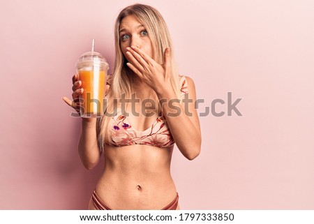 Young beautiful blonde woman wearing bikini drinking orange juice covering mouth with hand, shocked and afraid for mistake. surprised expression 