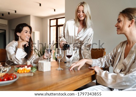 Beautiful cheerful girlfriends wearing dressing gowns celebrating together, drinking wine in the kitchen