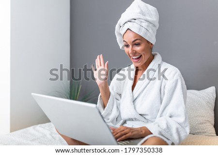 Beautiful young woman in white bathrobe in bedroom at home taking to her friends via video call . Smiling young woman lying on bed in bathrobe relaxing on the bed after bath and looking at her laptop