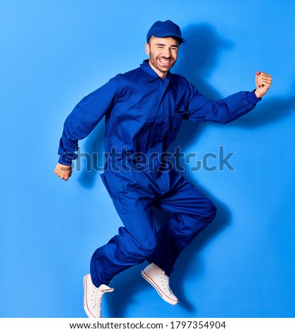 Young handsome hispanic man wearing painter uniform and cap smiling happy. Jumping with smile on face doig winner sign with fists up over isolated blue background