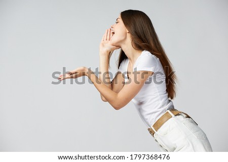 A woman in a white T-shirt and shorts shouts to the side on a light background and holds her hand 