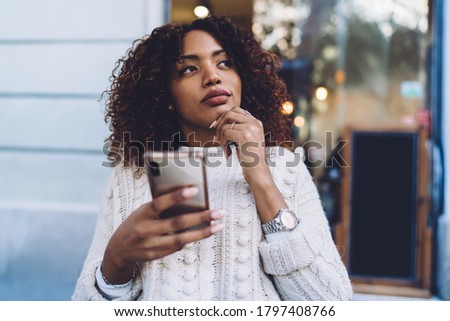 Pensive African American female in casual clothes sitting at wooden table and browsing mobile phone while resting in outdoor cafe