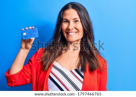 Young beautiful brunette businesswoman holding credit card over isolated blue background looking positive and happy standing and smiling with a confident smile showing teeth