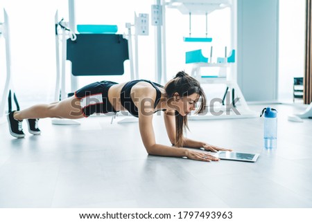 Side view of full body of young female in sportswear doing elbow plank and using tablet  on floor in modern gym