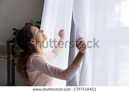 Happy young Caucasian woman open curtains breathe fresh air enjoy early morning sunshine, smiling relaxed millennial female welcome new sunny day, feel optimistic positive, stress free concept Royalty-Free Stock Photo #1797516451