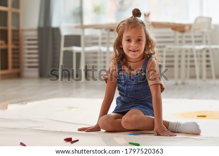 Full length portrait of cute little girl smiling at camera while sitting on floor in playroom and drawing, copy space