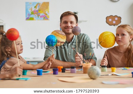 Portrait of smiling male teacher holding planet models and looking at camera while working with group of children in art and craft lesson, copy space