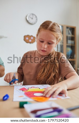 Vertical portrait of cute red haired girl cutting picture of space rocket while enjoying art and craft class in school or children development center