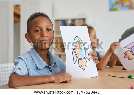 Portrait of cute African-American boy showing pictures of space rocket and smiling at camera while enjoying art class in pre school or children development center, copy space