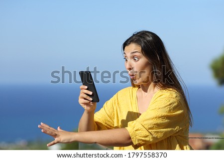 Surprised woman checks smart phone in a hotel balcony on the beach on summer vacation Royalty-Free Stock Photo #1797558370