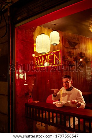 A European man eating Ramen in front of the storefront of a Japanese restaurant at night.