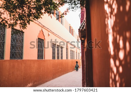 Woman in traditional arabic black cape and paranja carrying bag on her head on Marrakesh street. View from back. Red city, terracotta color background.