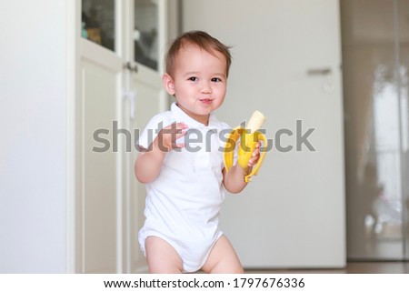 Happy baby boy eating banana and smiling. Mixed  race Asian-German infant self feeding fruit. Kid with healthy nutrition finger food fine motor development. Royalty-Free Stock Photo #1797676336