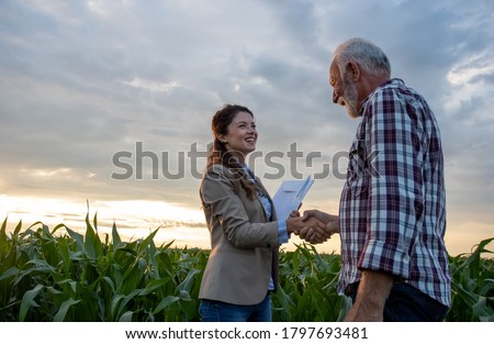 Senior man farmer shaking hands with young pretty woman with notebook in corn field. Insurance in agribusiness concept Royalty-Free Stock Photo #1797693481