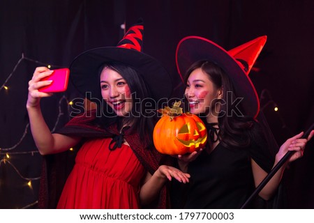 Two women taking photos with mobile phone at Halloween party. Selfie with mobile phone in the party. A woman dressed as a witch and a devil holding a pumpkin.