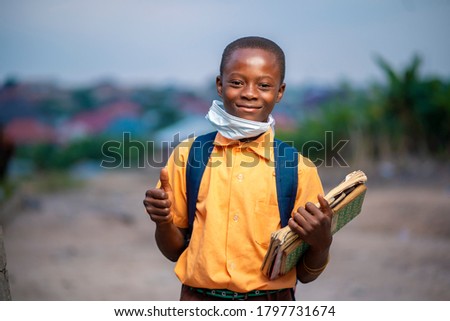 young African boy in uniform, with back pack and books in the hand-Black guy with face mask lowered at the chin with thumbs up sign-smiling  boy-school concept