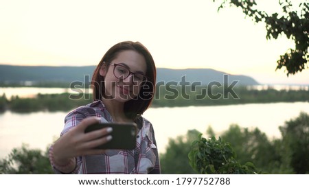 A young woman stands against the background of the river and mountains and takes pictures of herself.
