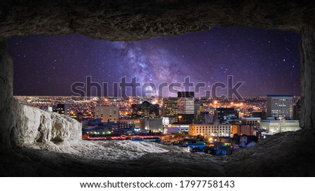 View from the inside of the cave to the night panorama of the city, buildings nicely lit by city lighting. On the horizon you can see a clear milky way in the night sky. 