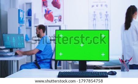 Dolly shot of computer with green screen in hospital cabinet with doctor and assistant working in the background.