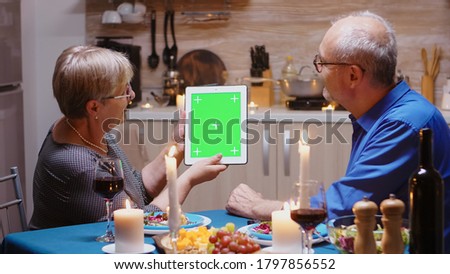 Old retired senior couple using green mock-up screen digital isolated tablet computer. Aged people looking at green screen template chroma key display sitting at the table in kitchen during dinner.