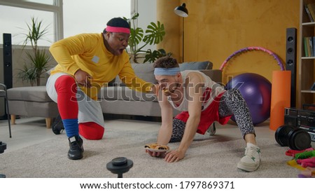 Hilarious fat afro-american instructor training weak unfit bearded man lifting up cookies plate like dumbbells. Fake fitness. Parody. Concept of fun, entertainment. Royalty-Free Stock Photo #1797869371