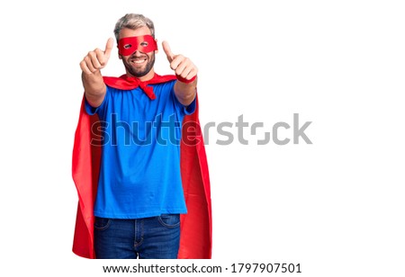 Young blond man wearing super hero custome approving doing positive gesture with hand, thumbs up smiling and happy for success. winner gesture. 