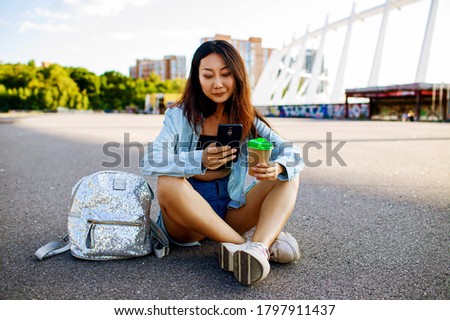 Young Asian Teen Girl With Backpack And Coffee Using Smartphone Outdoor.