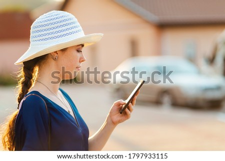 An elegant woman in a hat uses a smartphone. In the background, the house and car are blurred. Side view. The concept of social networking, business, and apps for phone