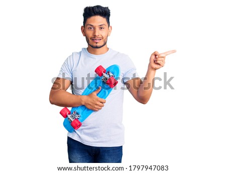 Handsome latin american young man holding skate smiling happy pointing with hand and finger to the side 