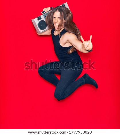 Young handsome man with long hair listening to music using vintage boom box. Jumping with smile on face doing heavy metal sign over isolated red background.