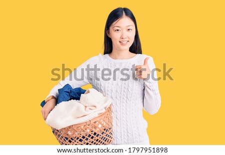 Young beautiful chinese woman holding laundry basket with clothes smiling happy and positive, thumb up doing excellent and approval sign 