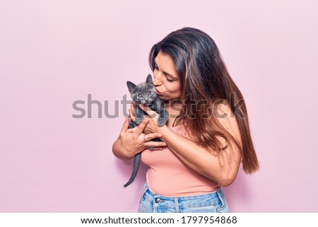 Young beautiful latin woman smilling happy. Standing with smile on face holding and kissing adorable cat over isolated pink background