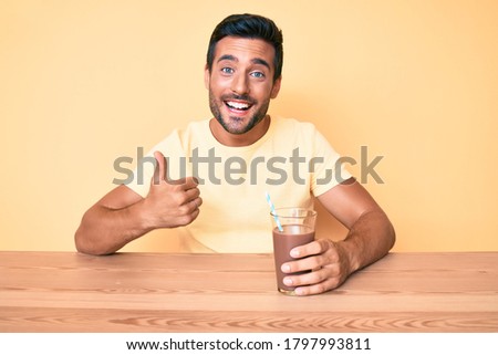 Young hispanic man sitting at the table drinking chocolate beverage smiling happy and positive, thumb up doing excellent and approval sign 