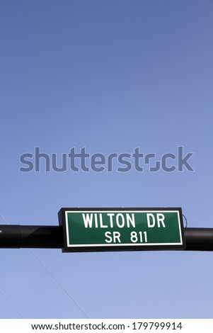 Rectangle white green and black street sign hanging from a pole on a sunny blue sky day in Wilton Manors, Florida. 
