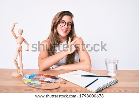 Beautiful young caucasian woman artist painter sitting on desk smiling cheerful pointing with hand and finger up to the side 