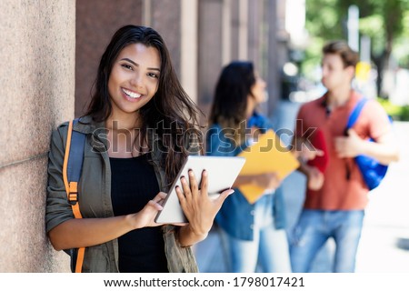 Laughing mexican student with tablet computer and friends in background in front of university building outdoor in summer in city Royalty-Free Stock Photo #1798017421