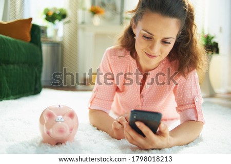 modern housewife in blouse and white pants with piggy bank searching for carpet cleaning service on a smartphone while laying on white carpet in the modern living room in sunny day.