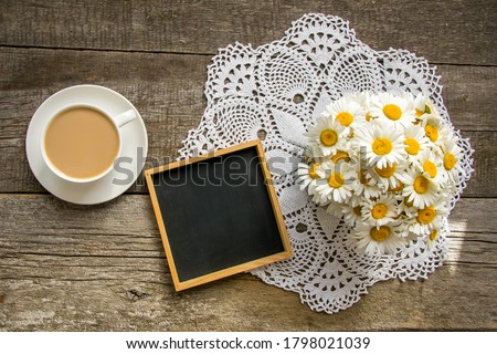 Top view of a black slate board in a wooden frame, a white cup with a saucer and coffee with milk, a bouquet of daisies on an old wooden background.