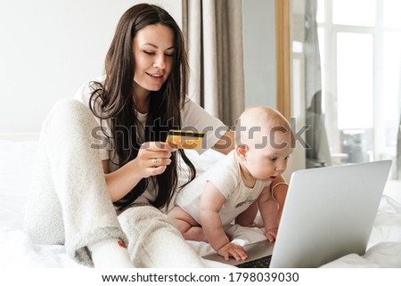 Photo of young happy caucasian mother with her baby using laptop and holding credit card on bed