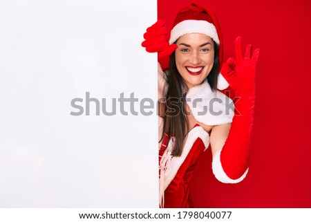 Young woman wearing santa claus costume holding blank empty banner doing ok sign with fingers, smiling friendly gesturing excellent symbol 